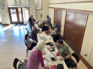 A group of people sit at two long, white tables. They are fixing a variety of items in front of them.