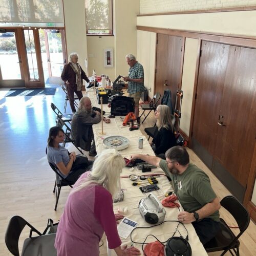 A group of people sit at two long, white tables. They are fixing a variety of items in front of them.