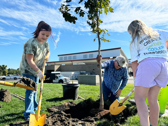 A girl in a green shirt and a girl in a white shirt are holding yellow shovels. They are scooping dirt into a hole where a man in a blue shirt is holding onto a tree they are planting. In the background, there is a school building.