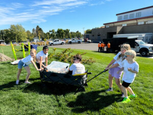 Kids in white t-shirts are pushing and pulling a black wagon. One kid is sitting in the wagon, along with a clear plastic tote box. In the background, there is a tree laying on its side. The kids are standing on green grass. The back of the picture is a parking lot and a school building. The sky is blue with wispy clouds.