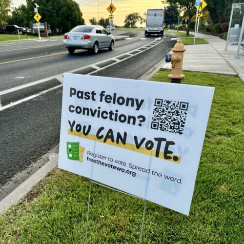 A white sign is staked in green grass next to a gray road with a silver car and white truck. There's a yellow fire hydrant in the back ground. The sign reads, "Past felony conviction? YOU CAN VOTE." There is a QR code on the sign.