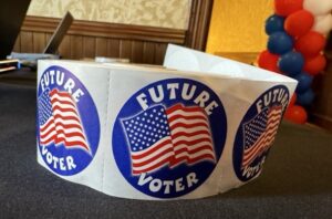 A roll of blue circle stickers with an American flg on it. The stickers read, "Future voter." The stickers are sitting on a gray table. In the background, there are red, blue and white balloons in a column.