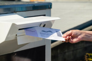 A hand holds a ballot. It is slipping the ballot into a white ballot drop box.