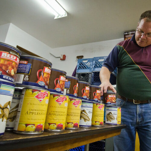 A man in a red and green shirt with blue jeans is standing next to two rows of canned food that's sitting on a table. He is in a small white room with fluorescent lights above him.