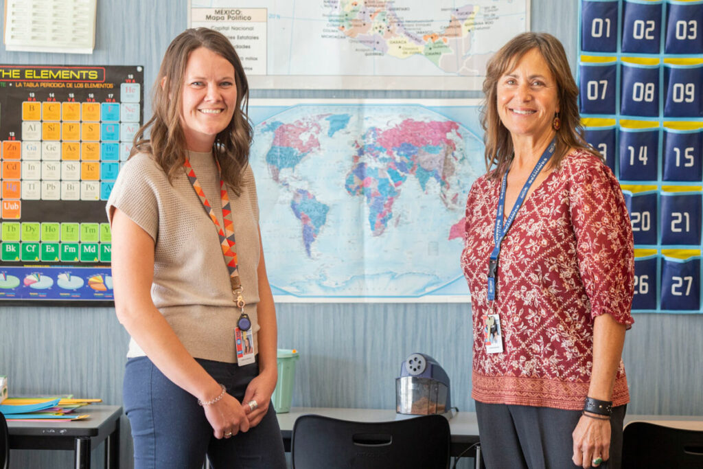 Two women stand in a classroom for a portrait. A map of the world is behind them.