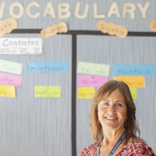 A woman wearing a red floral shirt stands in front of a wall with vocabulary words.