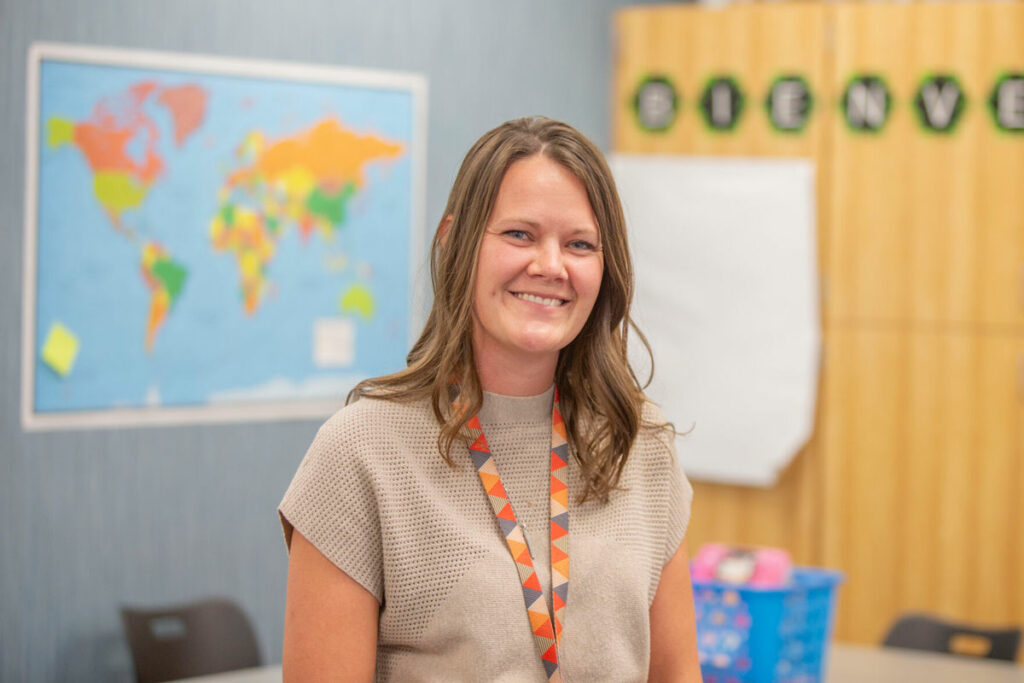 A woman wearing a light brown shirt and a lanyard stands in a classroom for a portrait. There is a map of the world behind her.